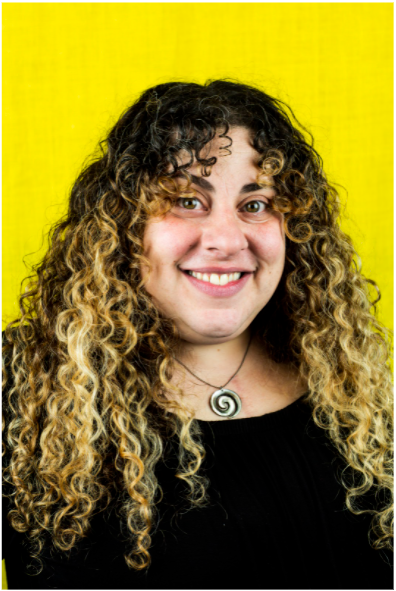 Oriana Leckert is a curly haired woman with a shell necklace and black top in front of a yellow background